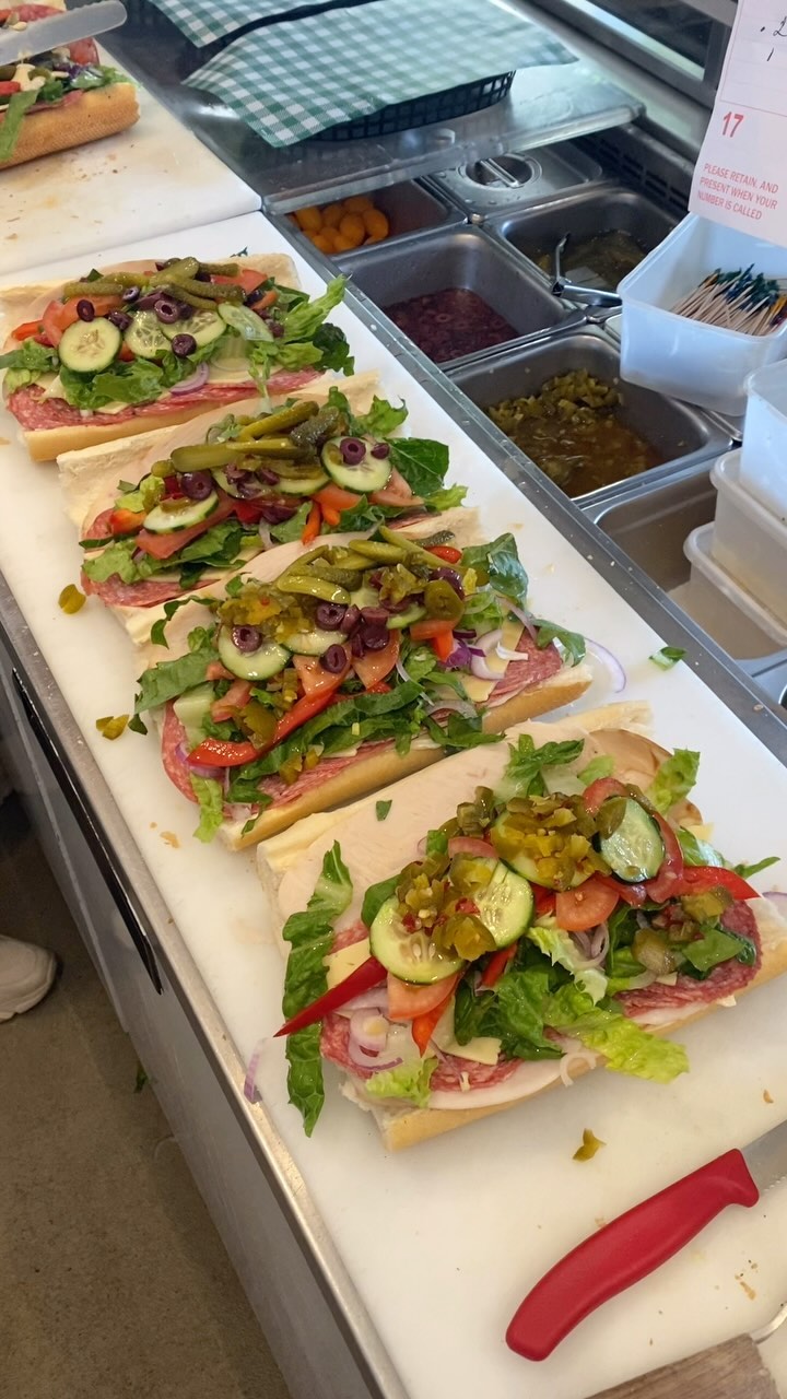 A row of subs being built on the prep bench with salami, salad, olives and jalapeños